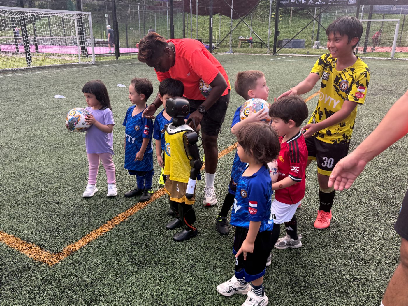 Young football players line up beside a Booster K1 humanoid robot wearing a team bib